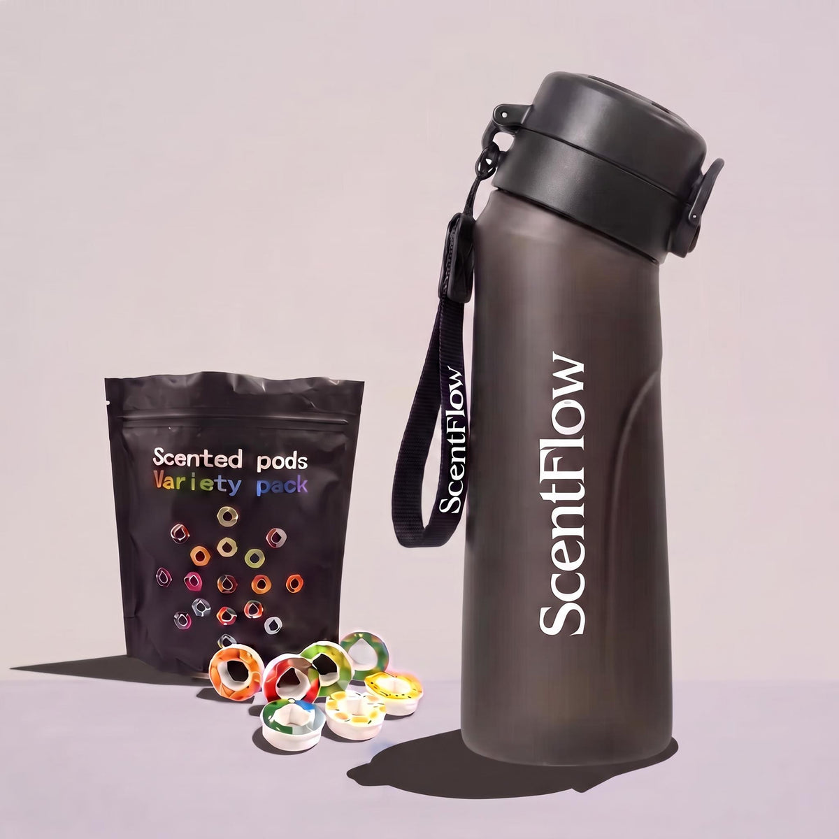 A matte dark gray ScentFlow insulated water bottle with a black lid and wrist strap stands next to a black ScentFlow pouch labeled 'Scented pods Variety pack' with several colorful pods scattered in front, on a light gray studio background.